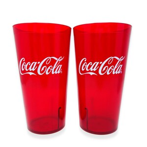 May include: Two translucent red plastic tumblers, each featuring the white Coca-Cola logo printed on the front. These cylindrical drinking glasses have a smooth finish and a slightly tapered shape, standing side by side against a plain white background.