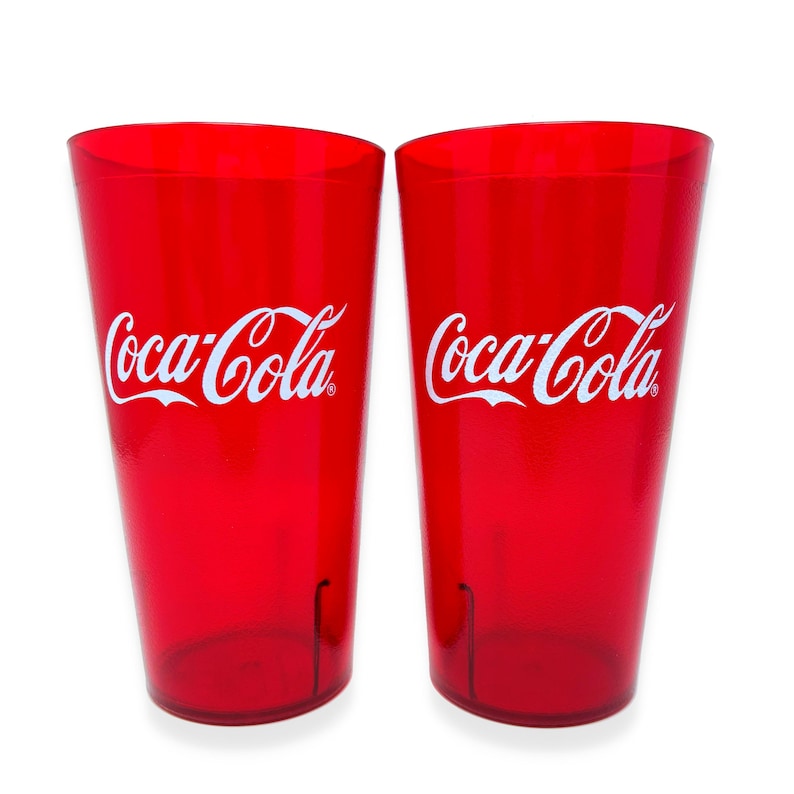 May include: Two translucent red plastic tumblers, each featuring the white Coca-Cola logo printed on the front. These cylindrical drinking glasses have a smooth finish and a slightly tapered shape, standing side by side against a plain white background.