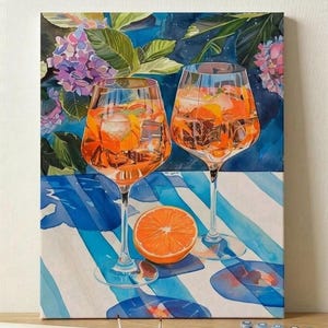 Op de afbeelding: Een canvas schilderij toont twee wijnglazen gevuld met een oranje drankje, ijs en sinaasappelschijfjes. Een doorgesneden sinaasappel ligt op een blauw-wit gestreept oppervlak. De achtergrond toont bloemen en bladeren.