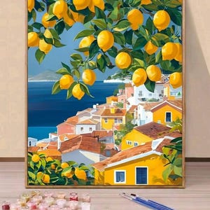 Op de afbeelding: Een ingelijst schilderij van een Italiaans kustdorp met gele gebouwen en oranje daken. Citroenboomtakken met gele vruchten hangen over het dorp. Het schilderij toont een blauwe zee en lucht.