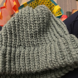 May include: A soft, chunky knit beanie in a muted green color. The beanie is folded over and has a ribbed texture.