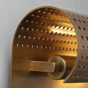 May include: A brass wall sconce with a perforated, curved shade and a cylindrical base. The light fixture has a warm, ambient glow.