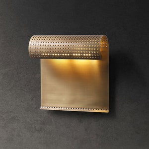 May include: A brass wall sconce with a perforated cylindrical shade and a rectangular backplate. The light is on and illuminates the wall behind it.