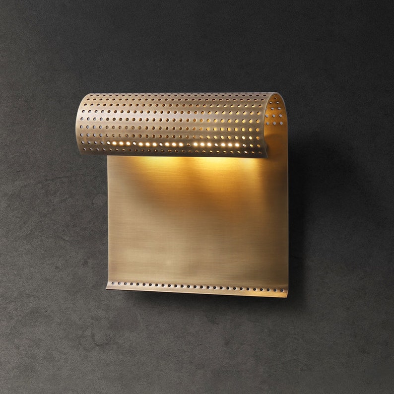 May include: A brass wall sconce with a perforated cylindrical shade and a rectangular backplate. The light is on and illuminates the wall behind it.