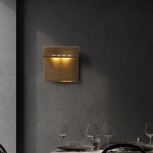 May include: A gold-toned wall sconce with a geometric pattern and a rectangular light fixture. The sconce is mounted on a dark gray wall.