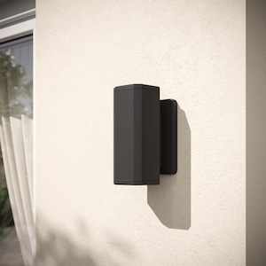 Könnte beinhalten: Eine schwarze Außenwandleuchte mit rechteckiger Form und leicht schrägem Design. Die Leuchte ist an einer weißen Wand montiert.