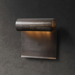May include: A silver wall sconce with a perforated metal shade and a rectangular metal back plate. The light is on and illuminating the back plate.