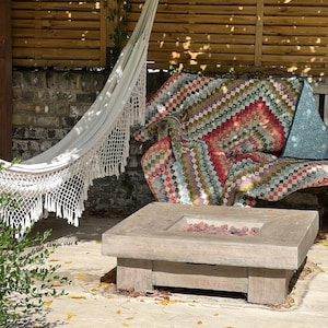 May include: A garden scene with a white fringed hammock, a wooden bench draped with a colourful quilt, and a square concrete fire pit filled with red stones. The setting is a patio with a wooden fence and brick wall.