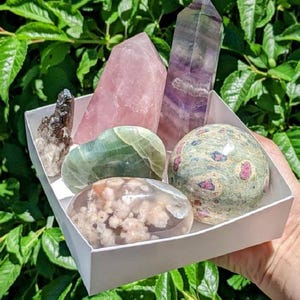 Pode incluir: Uma caixa branca cheia de seis tipos diferentes de cristais. Os cristais são de várias cores, incluindo rosa, verde, roxo e marrom. A caixa está sentada em uma mão.