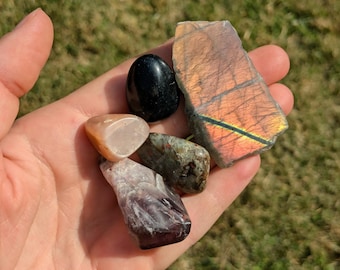 May include: A collection of five natural stones including a smooth black polished stone, a large flat piece of labradorite with orange and gold iridescence, a pale peach stone, a mottled green and brown stone, and a dark purple amethyst crystal.