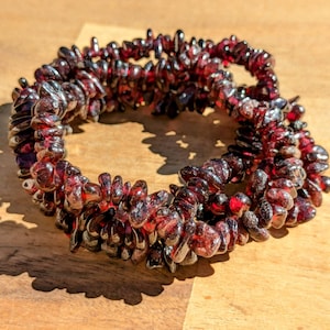 May include: A bracelet made of small, irregularly shaped, deep red gemstones. The stones are strung together to form a circular shape. The bracelet is resting on a light brown wooden surface, with shadows cast across the wood.