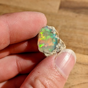 Op de afbeelding: Een ruwe, natuurlijke opaal edelsteen met een groene, blauwe en oranje iriserende glans. De opaal is ingebed in een matrix van grijs gesteente.