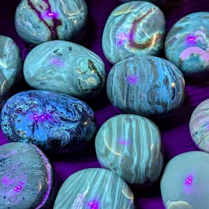 May include: A collection of 11 smooth, polished stones with a blue and grey marbled pattern. The stones are illuminated with a UV light, revealing a bright pink fluorescence.