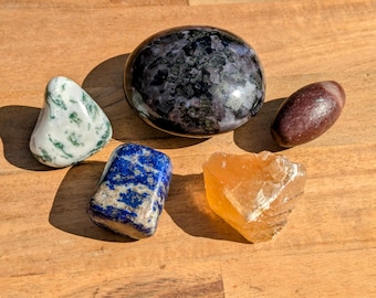 May include: A collection of five polished stones, including a green and white agate, a blue lapis lazuli, a brown and white stone, a yellow calcite, and a black and white speckled stone.