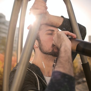 Puede incluir: Un hombre con una sudadera con capucha negra y una camiseta blanca sostiene una botella de agua de acero inoxidable y bebe de ella. Se apoya en una estructura metálica.