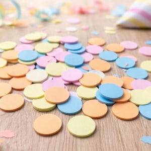 Puede incluir: Confeti colorido en tonos naranja, amarillo, rosa y azul esparcido sobre una superficie de madera. Serpentinas y un gorro de fiesta son visibles en el fondo, sugiriendo un tema festivo.