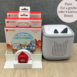 Może przedstawiać: Biały stojak ekspozycyjny z trzema czerwono-białymi pudełkami Tonie Play Game, czerwoną figurką Tonie i szarą Toniebox. Pudełka przedstawiają postacie z kreskówek i tekst "LALALINOS DEIN TAG IN BUMBALOO". Toniebox ma czarne akcenty w kształcie uszu.