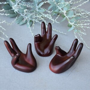 May include: Three wooden hand-shaped ring holders. The holders are a dark reddish-brown colour and are arranged in a semi-circle on a grey surface.