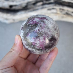 May include: A spherical gemstone, displaying a mix of white, grey, and pink hues. The stone has a cloudy, marbled appearance. It is held in a hand. The background features a light grey surface and a piece of driftwood.
