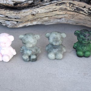 May include: Four small, carved stone teddy bear figurines in varying colours: light pink, two shades of grey, and dark green. Each bear has a heart-shaped detail on its chest. The bears are displayed on a grey surface with a piece of driftwood in the background.