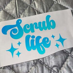 Op de afbeelding: Een blauwe en witte vinyl sticker met de tekst "Scrub Life" in een gestileerd lettertype. De sticker heeft drie kleine, glinsterende sterren.