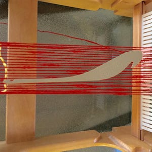 May include: A wooden weaving loom with red yarn stretched across. A light-colored shuttle is in the center, and a white and clear plastic reed is on the right side. The loom is partially assembled, showing the frame and components.