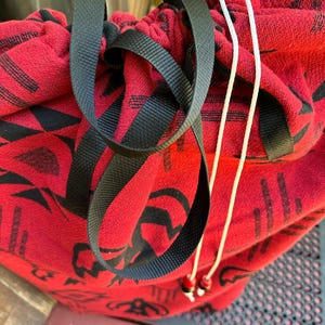 May include: A red drawstring bag with black and white geometric patterns. The bag is tied with a black drawstring and a white cord.