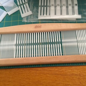 May include: A wooden Ashford rigid heddle loom with white plastic heddles. The loom is on a green cutting mat.