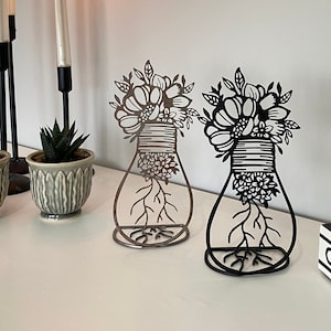 Puede incluir: Dos esculturas florales de metal negro con un diseño de jarrón y raíces. Las esculturas están sobre una superficie blanca con dos pequeñas plantas en maceta y un candelero con tres velas.