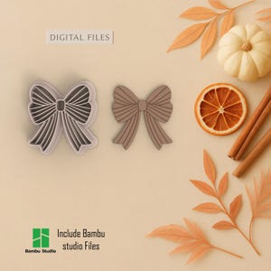 May include: Two bow-shaped cookie cutters, one with a white outline and one solid brown, sit on a beige surface. The words "DIGITAL FILES" are above the cutters. A small pumpkin, dried orange slice, and cinnamon sticks are also present.
