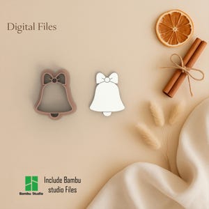 Puede incluir: Dos cortadores de galletas en forma de campana, uno con contorno marrón claro y otro blanco, sobre una superficie beige. La imagen incluye una rodaja de naranja seca, palitos de canela y el texto "Digital Files" e "Include Bambu studio Files."