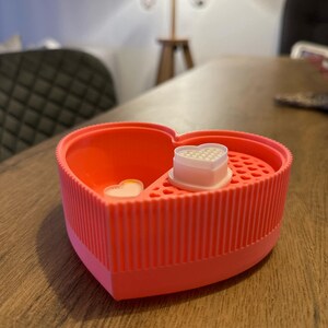 May include: A pink heart-shaped silicone container with a white heart-shaped mold inside. The mold has small holes in it.