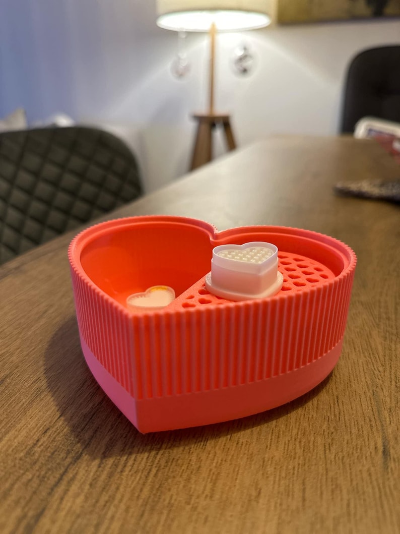 May include: A pink heart-shaped silicone container with a white heart-shaped mold inside. The mold has small holes in it.