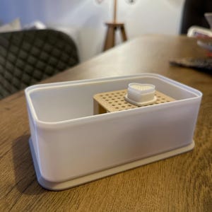 May include: A white plastic container with a beige insert and a white heart-shaped lid. The container is designed for storing small items.