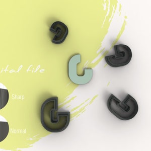 Puede incluir: Cinco cortadores de galletas en forma de la letra "G" con un doble contorno. Cuatro son negros y uno es azul claro. Los cortadores de galletas están sobre una superficie blanca con un fondo amarillo.