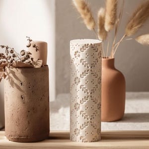 Op de afbeelding: Een hoge, cilindrische crèmekleurige kandelaar met een bloemen- en bladerpatroon. De kandelaar staat naast een bruine vaas met gedroogde planten en een getextureerde bruine betonnen kandelaar.