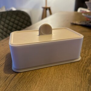 May include: A white rectangular box with a beige lid and a small handle on top. The box is sitting on a wooden surface.