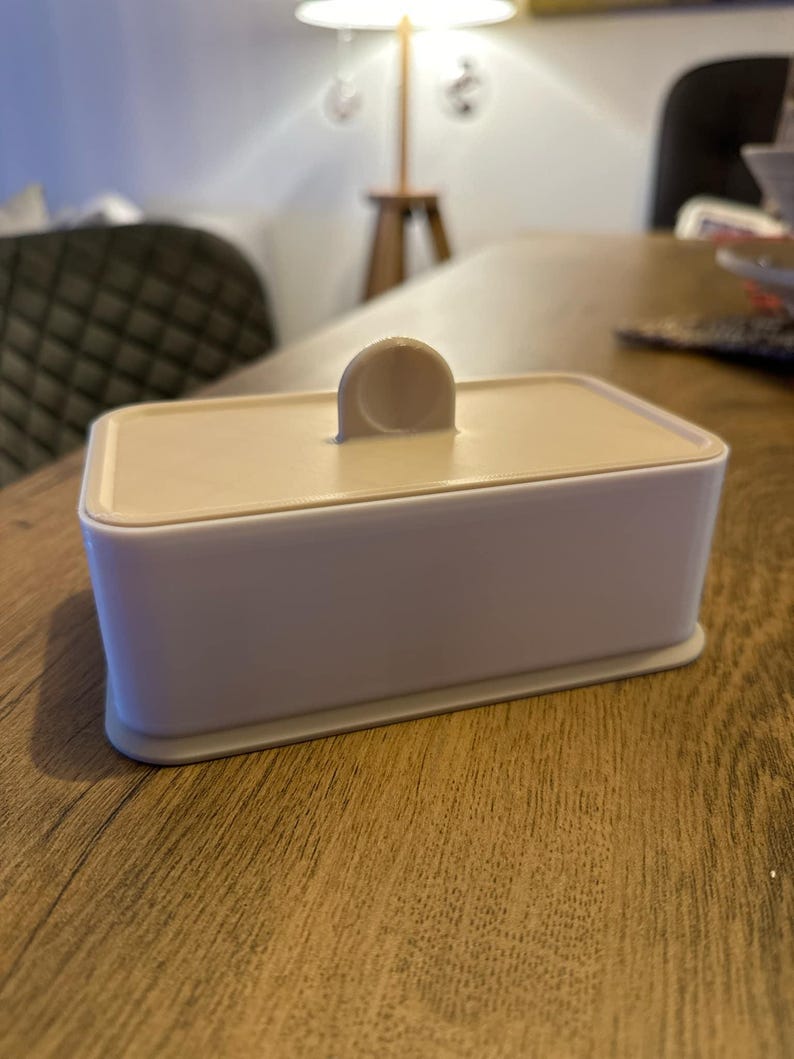 May include: A white rectangular box with a beige lid and a small handle on top. The box is sitting on a wooden surface.