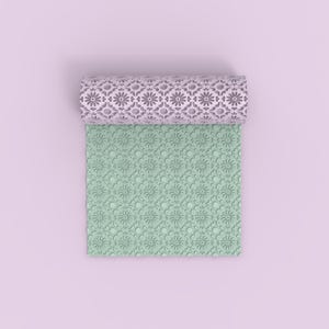 May include: A rolled, pale purple patterned rolling pin sits above a light green, floral-patterned mat. The rolling pin and mat feature a repeating floral design. The background is a pale purple.