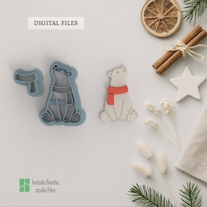 May include: A white polar bear cookie cutter with a red scarf is next to a blue cookie cutter of a polar bear wearing a scarf. The image also includes a dried orange slice, cinnamon sticks, and a white star.