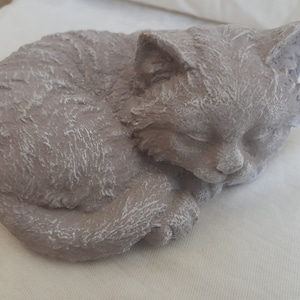 May include: A curled-up cat ornament, coloured in muted grey with white highlights. The cat's eyes are closed, suggesting sleep. The sculpture has a textured finish, giving it a rustic look. A decorative item for cat enthusiasts.
