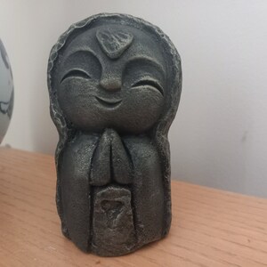 May include: A small, grey statue of a smiling figure with hands clasped in prayer. The statue has a textured surface and is set against a neutral background. The figure's head is adorned with a leaf-shaped detail.