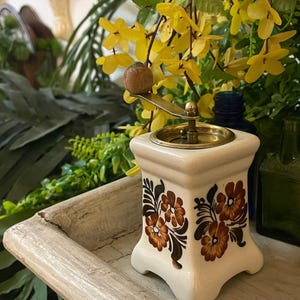 Könnte beinhalten: Cremefarbene Kaffeemühle aus Keramik mit Holzgriff und Messingakzenten. Die Mühle hat braune Blumenmuster an den Seiten. Sie steht auf einem Holztablett, mit gelben Blumen im Hintergrund. Ein Vintage-Küchenartikel.