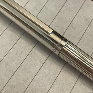 Puede incluir: Primer plano de un bol&iacute;grafo plateado con una superficie brillante y reflectante. El bol&iacute;grafo tiene un clip rectangular y un dise&ntilde;o a rayas en el cuerpo inferior. La marca "S.T. Dupont" est&aacute; grabada en el bol&iacute;grafo.