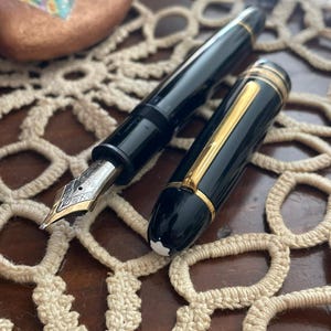 May include: Two black fountain pens with gold accents are displayed on a beige crocheted doily. One pen has a gold nib with intricate designs. The other pen has a gold band and a white emblem.