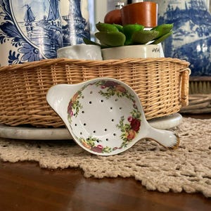 May include: A white porcelain tea strainer with a floral design in red, yellow, and green. The strainer has a handle and small holes for straining tea leaves. The rim is gold. It sits on a woven basket.