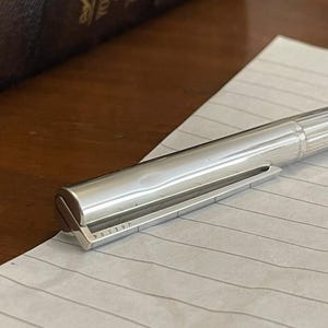 Puede incluir: Un bol&iacute;grafo plateado descansa sobre papel rayado. El bol&iacute;grafo tiene un dise&ntilde;o met&aacute;lico elegante con un cuerpo acanalado y una punta plana y angular. El n&uacute;mero de serie del bol&iacute;grafo, "J03904", es visible en la punta. Un libro marr&oacute;n oscuro est&aacute; en el fondo.