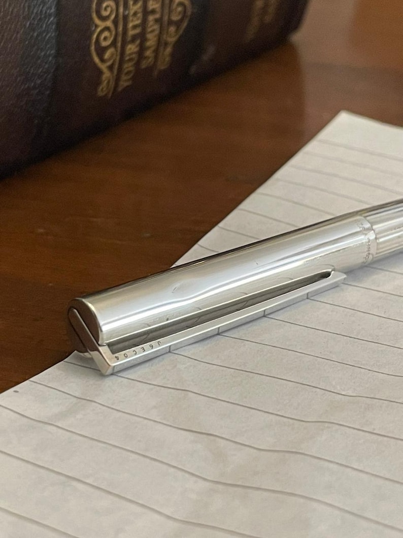 Puede incluir: Un bol&iacute;grafo plateado descansa sobre papel rayado. El bol&iacute;grafo tiene un dise&ntilde;o met&aacute;lico elegante con un cuerpo acanalado y una punta plana y angular. El n&uacute;mero de serie del bol&iacute;grafo, "J03904", es visible en la punta. Un libro marr&oacute;n oscuro est&aacute; en el fondo.