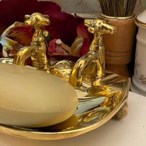 May include: A gold-toned metal soap dish shaped like a miniature bathtub with two faucet handles at one end. A smooth, oval-shaped beige bar of soap rests inside the basin of the dish. The metallic surface is reflective and polished.
