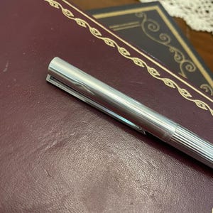 Puede incluir: Un bol&iacute;grafo plateado con clip descansa sobre un libro encuadernado en cuero burdeos. El bol&iacute;grafo tiene un dise&ntilde;o acanalado en la parte inferior del cuerpo y un acabado met&aacute;lico brillante. El libro tiene adornos dorados.